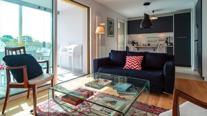 Un salon moderne avec une table basse en verre, un canapé bleu foncé agrémenté d’un coussin rouge et blanc, et une chaise en bois. On aperçoit une cuisine à l’arrière-plan, avec des meubles sombres et un luminaire suspendu.