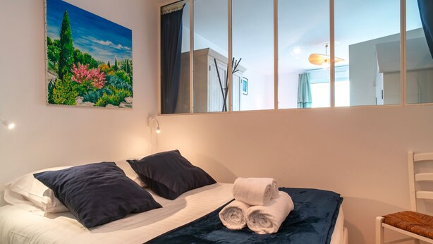 Un lit bien fait, avec une literie bleu foncé, un tableau représentant un jardin accroché au mur et une grande fenêtre donnant sur l’extérieur.
