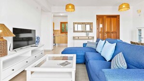 Een moderne woonkamer met een blauwe bank, een witte salontafel en een televisie op een witte kast. Achtergrond toont wilgentreinen lampen en een houten deur.