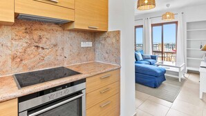 Een moderne keuken met granieten aanrechtbladen, een ingebouwde oven en houten kastjes. Grenzend aan de keuken bevindt zich een woonkamer met een blauwe bank, een salontafel en een balkon met uitzicht.