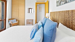 Een hotelkamer met een groot bed, een rieten stoel, een houten garderobe, een klein houten kistje, en een deur naar een andere kamer.