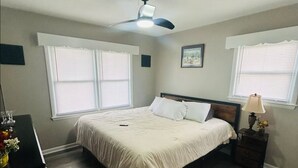 A bedroom with a bed, a ceiling fan, a nightstand with a lamp, and two windows with blinds. - El Rancho Powell (Goldsboro)