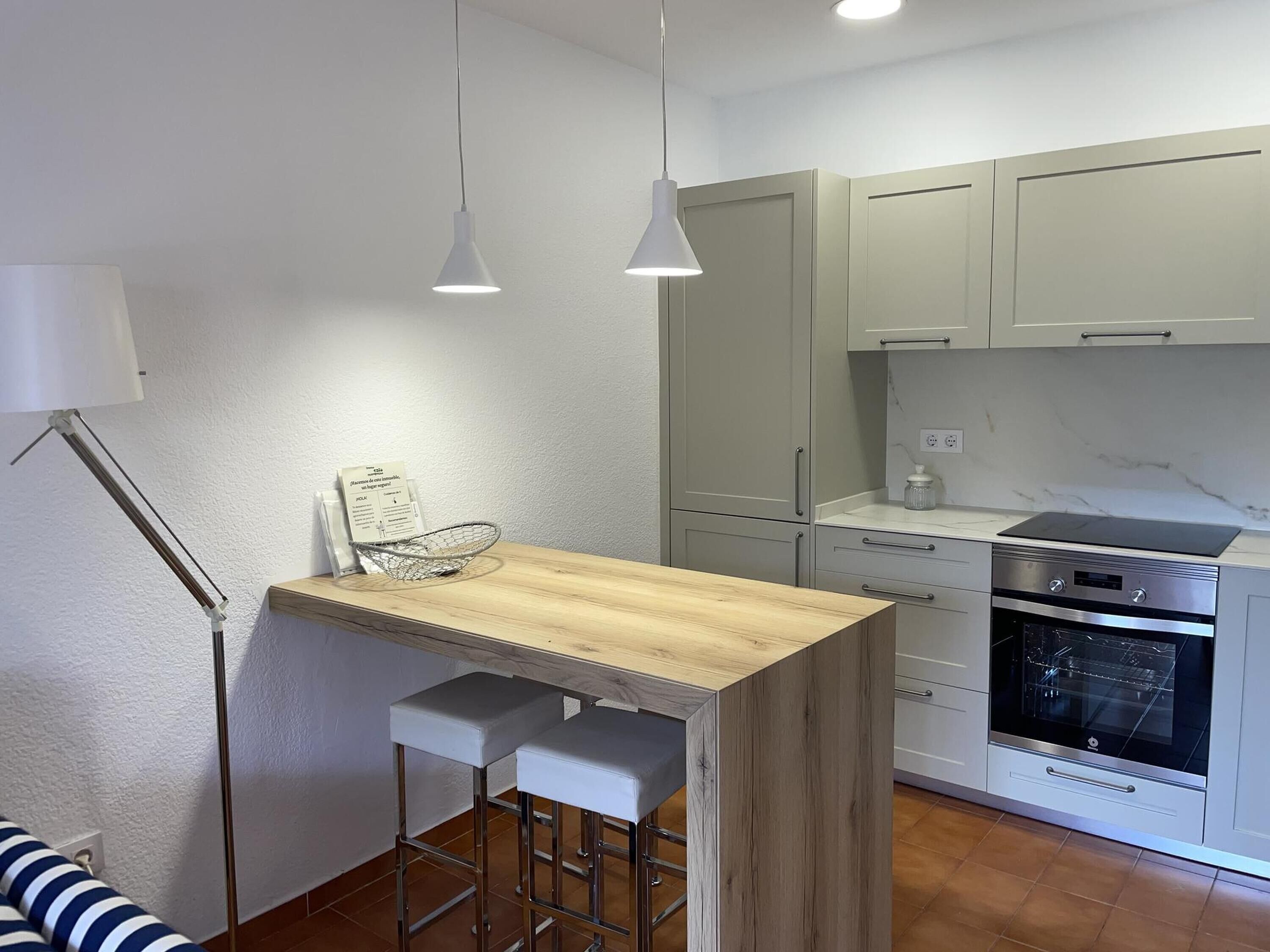 Eine kompakte Küche mit einer Holz-Kochinsel, zwei Barhocker, einem eingebauten Backofen und einer Pendelleuchte.