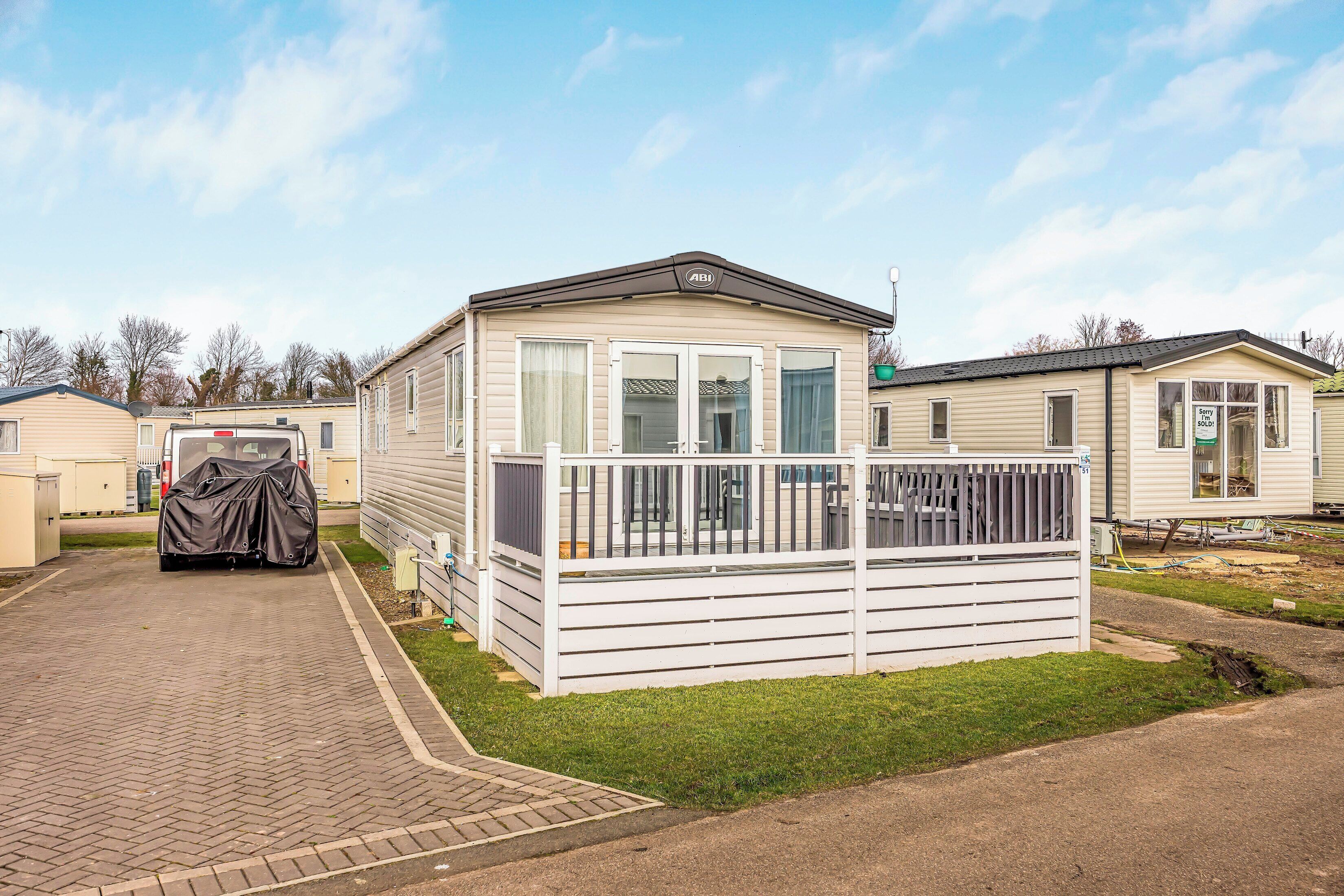 A mobile home with a covered vehicle in front, a fenced deck, and a clear sky.
