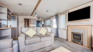 A compact living area with a sofa, a flat-screen TV, a fireplace, and a kitchenette. - Charming Home in Lakeside Park - Pass The Keys (Chichester)