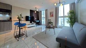 Una sala moderna con un sofá gris, una televisión, una zona de comedor con sillas azules y una cocina con horno empotrado.