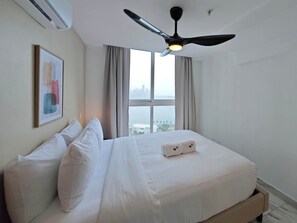 Una habitación de hotel con cama, ventilador de techo, ventana con cortinas y vista panorámica de la ciudad.