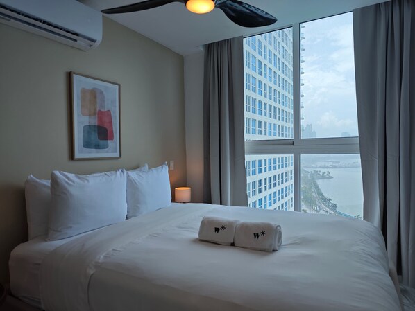 A hotel room with a large bed, a ceiling fan, a window with a city view, and a framed abstract painting on the wall. - w* | Sunlit 1BR on Avenida Balboa w/ Pool (Panamá)