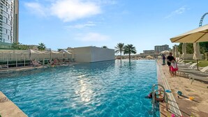Una piscina con agua cristalina azul, rodeada de sillas de descanso y sombrillas, con un edificio alto al fondo.
