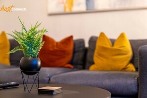 A modern living room with a grey sofa, yellow and orange cushions, a small black table with a potted plant and a stack of books. - Cosy 2-Bed House | Easy Edinburgh Access | Sleeps 4 (Whitburn)