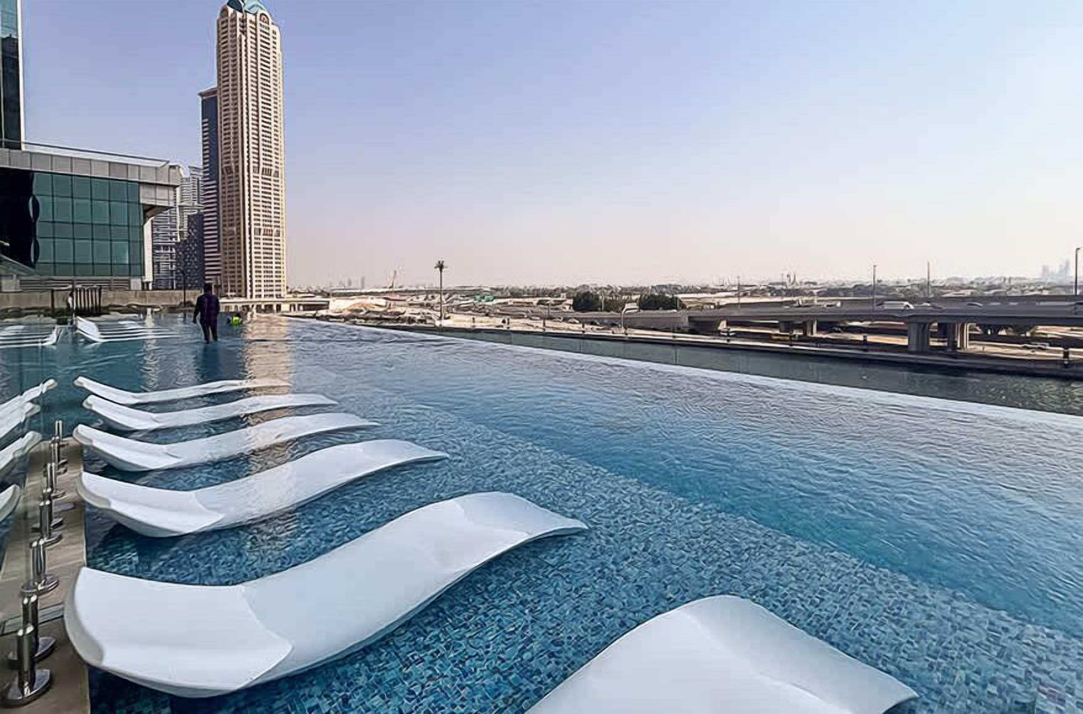 En infinitypool med vita, flytande solstolar, utsikt över en stadssiluett och en bro i bakgrunden.