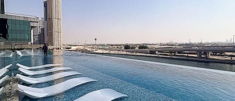 An infinity pool with white floating loungers, a view of a cityscape, and a bridge in the background.