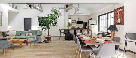 Un salon moderne avec un canapé, des fauteuils, une table à manger dressée pour six personnes et une cuisine en arrière-plan.