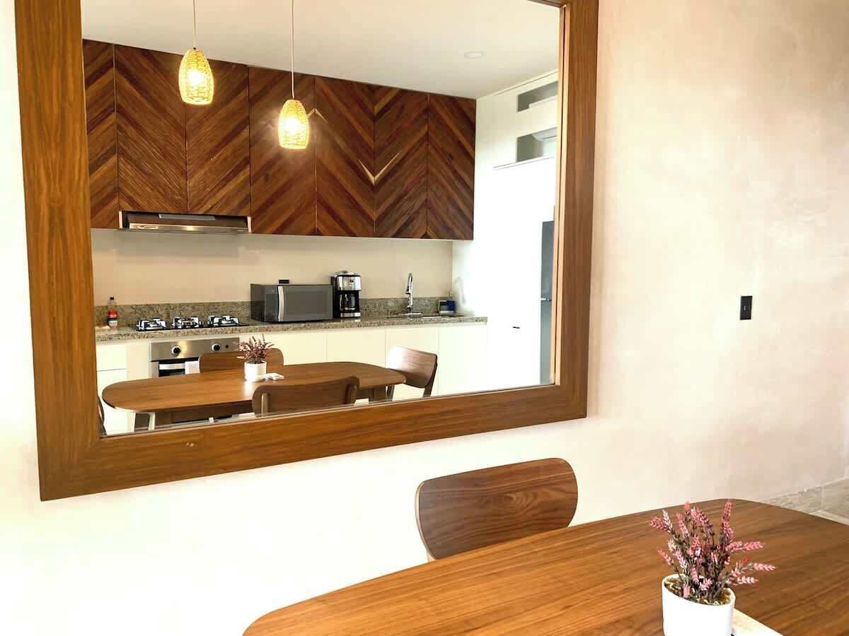 A dining area with a wooden table and chairs, a large mirror reflecting the kitchen, and pendant lighting.