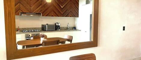 A dining area with a wooden table and chairs, a large mirror reflecting the kitchen, and pendant lighting.
