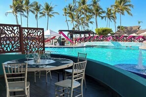 A poolside area with a table and chairs, surrounded by palm trees and a clear blue sky. - Ocean view, beach access and amenities! (Puerto Vallarta)