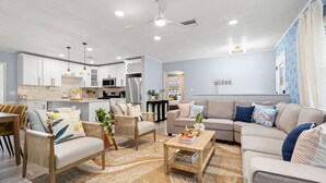 Un salon moderne avec un canapé, des fauteuils et une table basse. On aperçoit à l’arrière-plan un coin cuisine avec des placards et des appareils électroménagers.