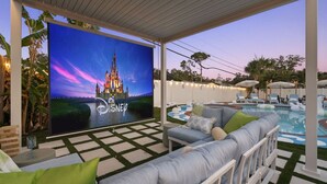 Un espace extérieur abrité doté d’un grand écran sur le thème de Disney, d’une piscine, de chaises longues et d’un coin salon avec des coussins.