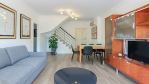 Ein modernes Wohnzimmer mit einem grauen Sofa, einem runden Couchtisch, einem Essbereich mit Holztisch und -stühlen, einer Treppe und einem Fernseher auf einer Holzkommode.