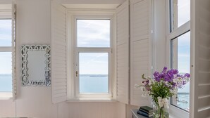 A room with a bay window, a view of the sea, a mirror, and a vase with flowers.