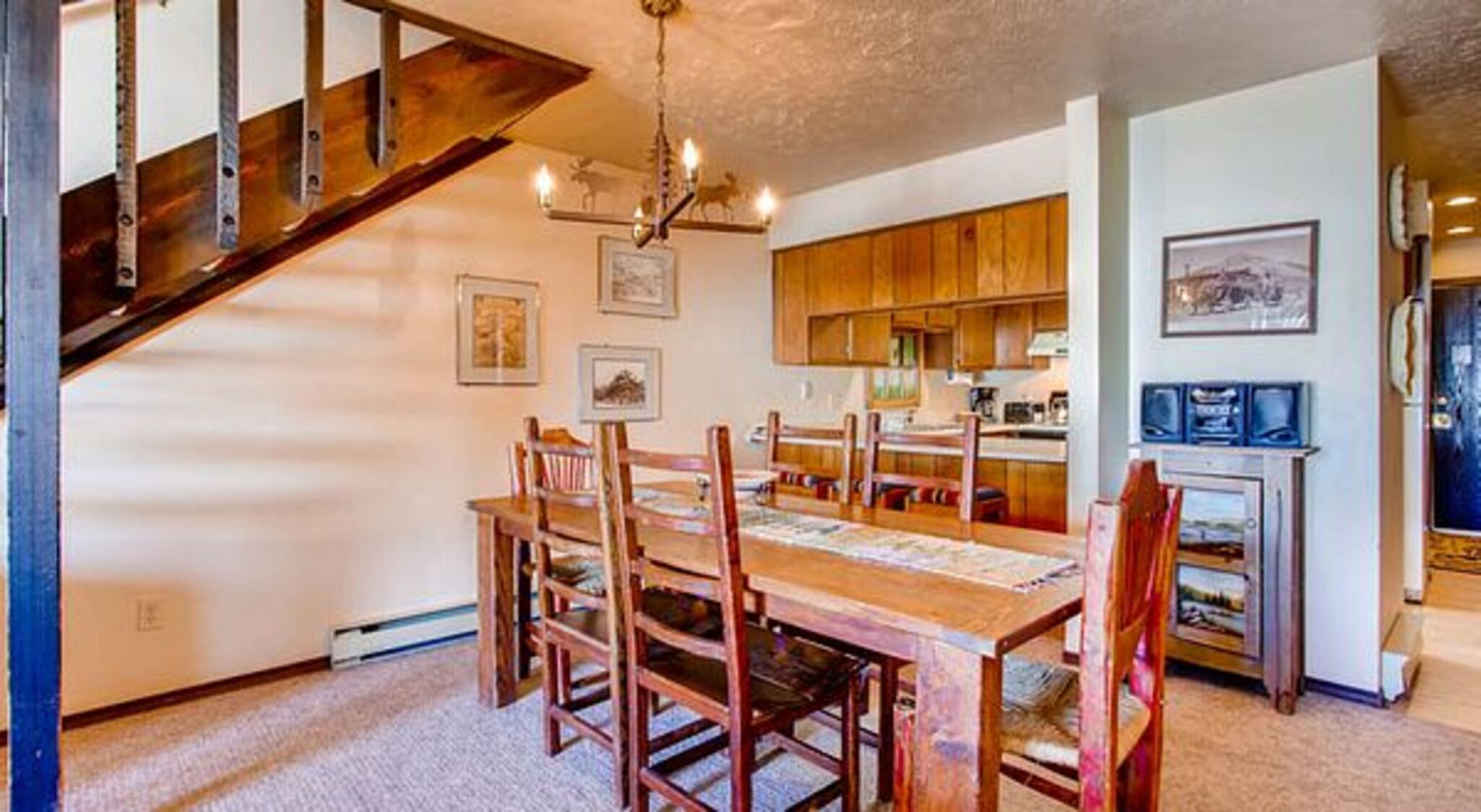 A dining area with a wooden table and chairs, a kitchen in the background, and a staircase leading to another level.
