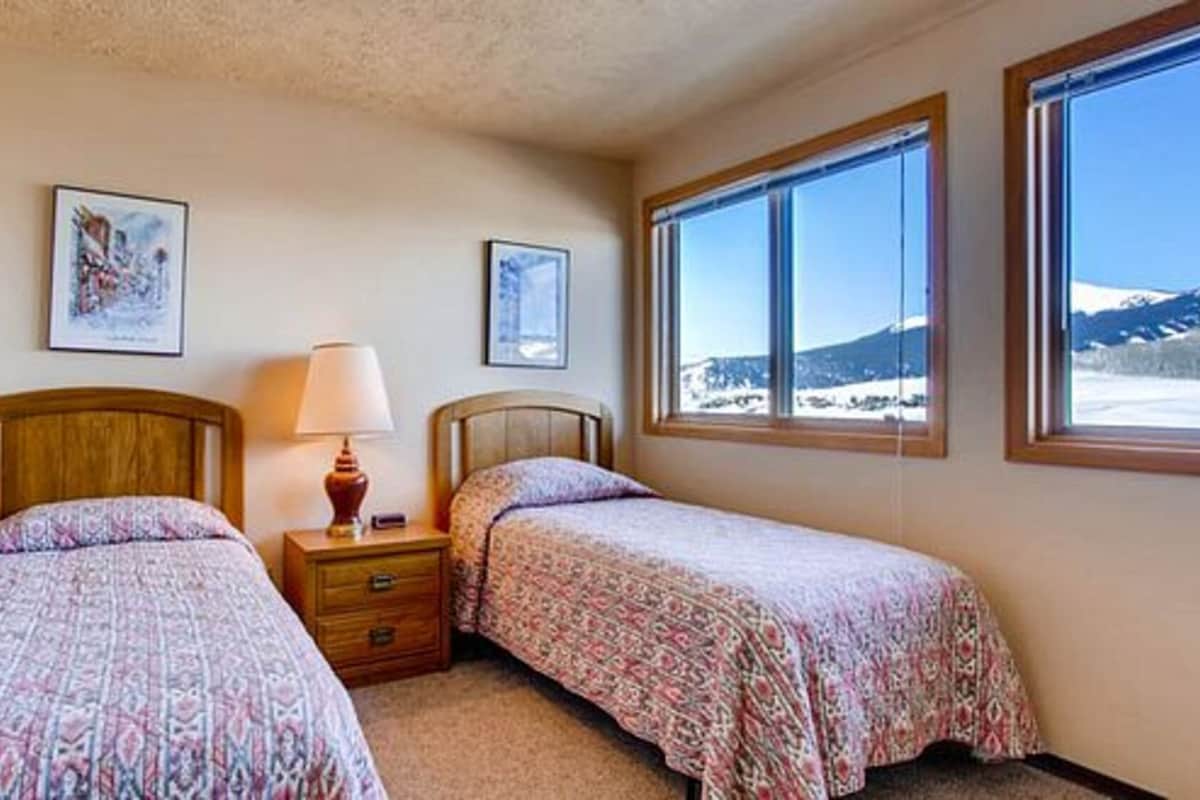 A bedroom with two beds, a nightstand with a lamp, and a window with a mountain view.