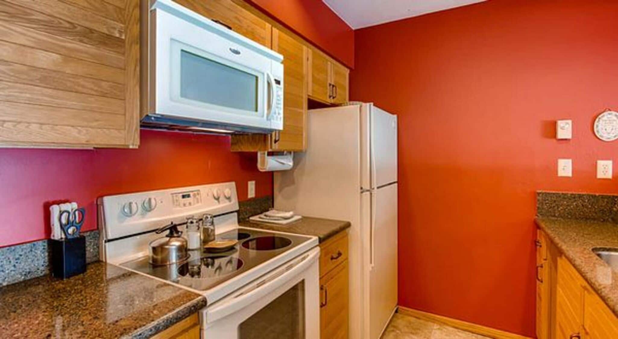 A kitchen with a white stove, oven, microwave, and refrigerator, wooden cabinets, and a red wall.
