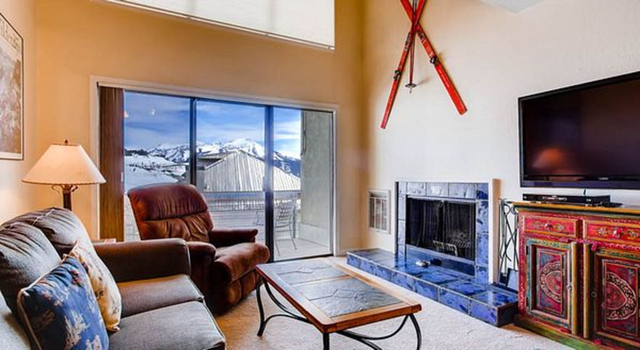 A living room with a sofa, a coffee table, a fireplace, a television, and a view of a snowy mountain through large windows.