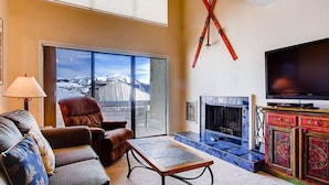 A living room with a sofa, a coffee table, a fireplace, a television, and a view of a snowy mountain through large windows. - Crested Butte Mountain Resort Properties by Vail (Crested Butte)