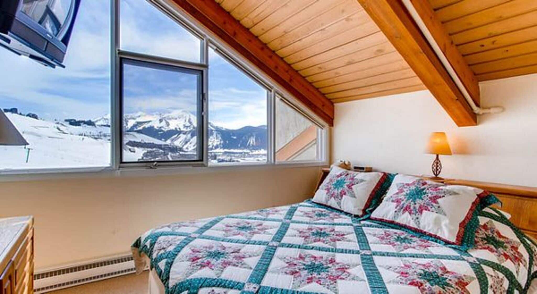 A bedroom with a large window offering a mountain view, a bed with a floral quilt, and a wooden ceiling.