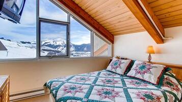 A bedroom with a large window offering a mountain view, a bed with a floral quilt, and a wooden ceiling.