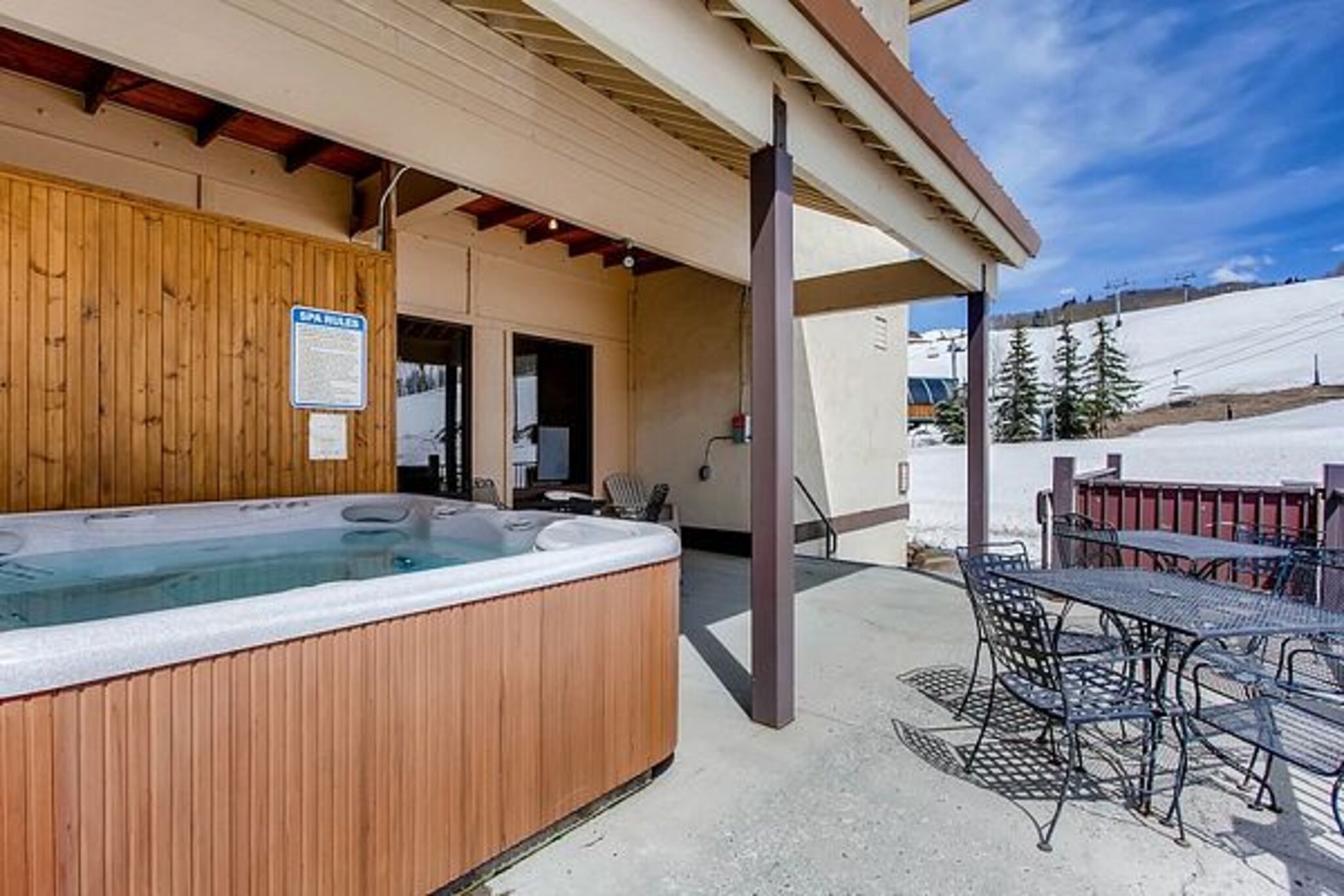 A hot tub with a wooden exterior, a covered patio area with metal chairs, and a view of a snowy landscape.