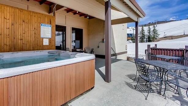 A hot tub with a wooden exterior, a covered patio area with metal chairs, and a view of a snowy landscape. - Crested Butte Mountain Resort Properties by Vail (Crested Butte)