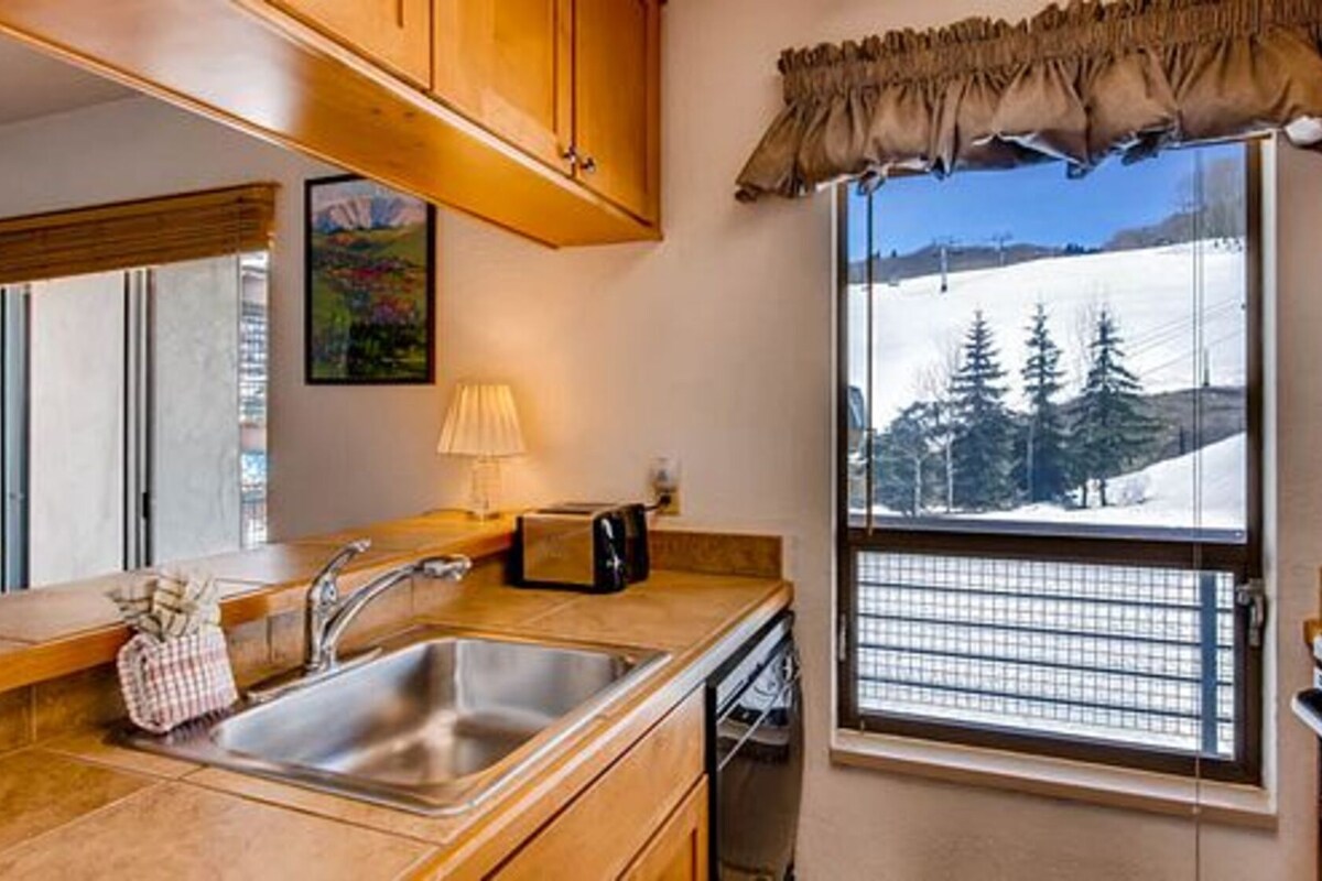 A kitchen with wooden cabinets, a sink, a window with a view of trees, and a painting on the wall.