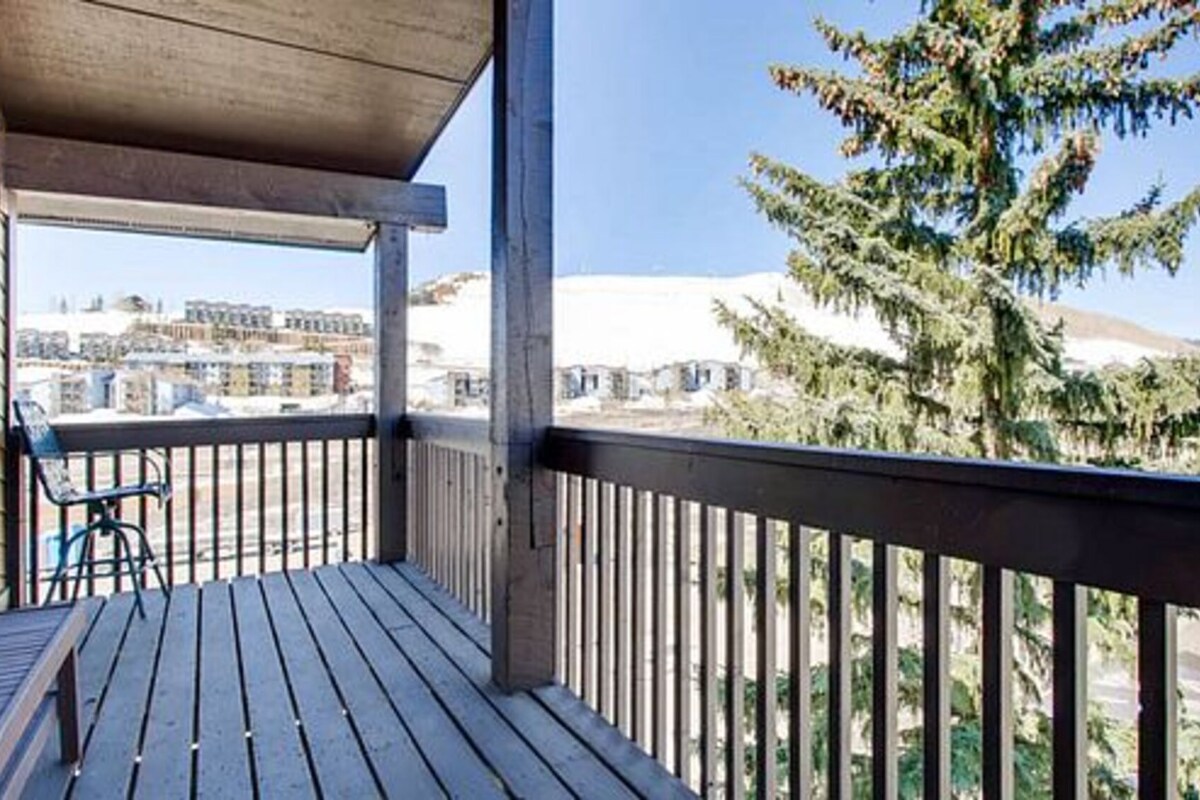 A wooden balcony with a view of buildings and a coniferous tree.