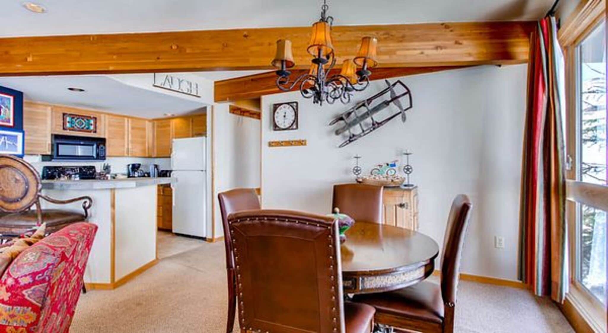 A dining area with a wooden table and chairs, a chandelier, and a kitchen in the background.
