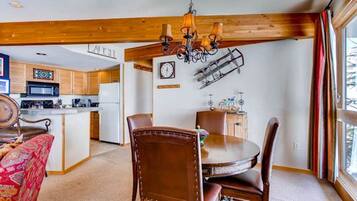 A dining area with a wooden table and chairs, a chandelier, and a kitchen in the background.