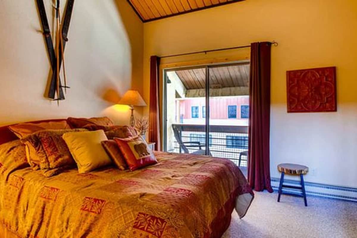 Un dormitorio con una cama grande, una mesita de noche, una lámpara, un taburete pequeño y vistas al balcón exterior.