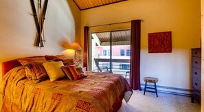 A bedroom with a large bed, a nightstand, a lamp, a small stool, and a view of the outside balcony. - Crested Butte Mountain Resort Properties by Vail (Crested Butte)