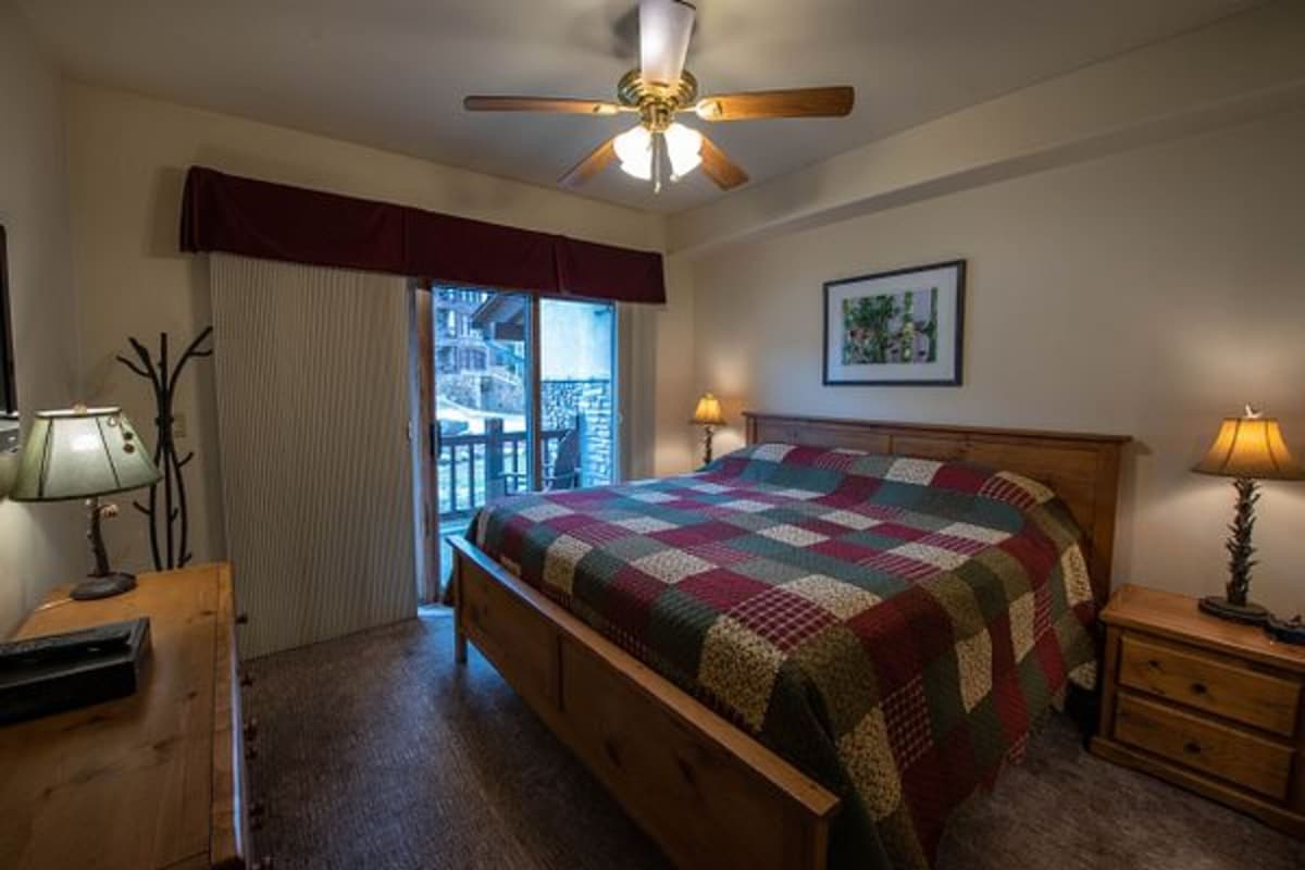 A bedroom with a wooden bed, a ceiling fan, a window with curtains, a nightstand with a lamp, and a framed picture on the wall.