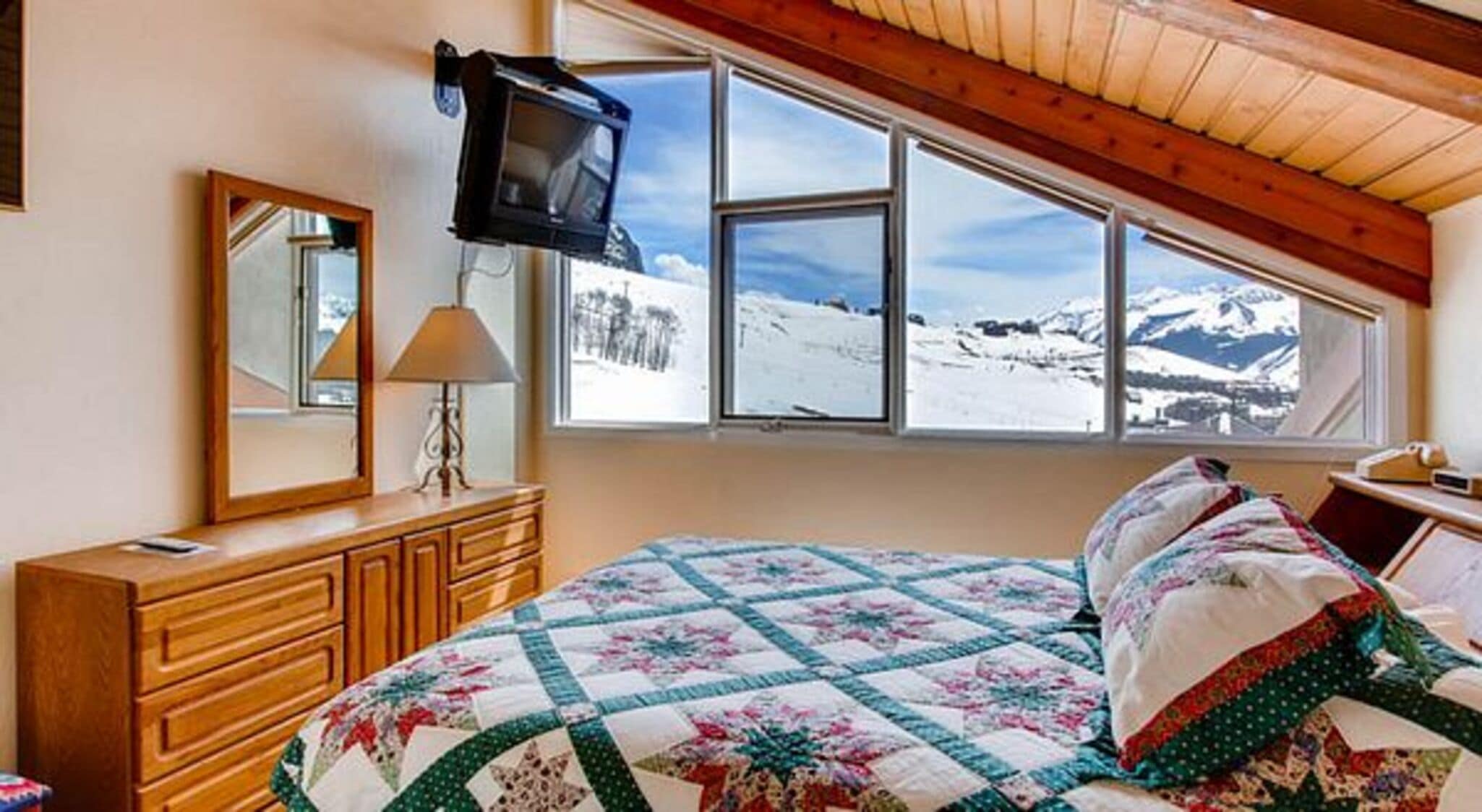 A bedroom with a bed, a dresser, a TV mounted on the wall, and a window with a snowy mountain view.