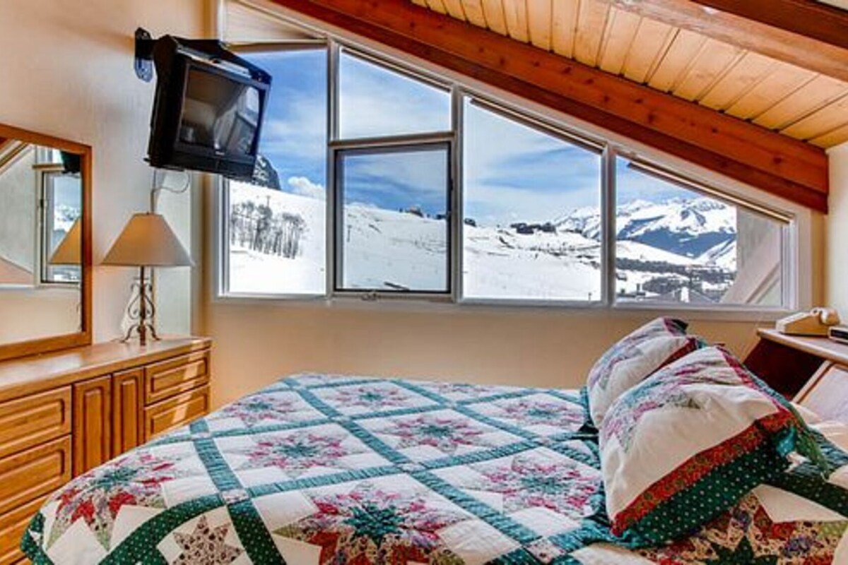 A bedroom with a bed, a dresser, a TV mounted on the wall, and a window with a snowy mountain view.