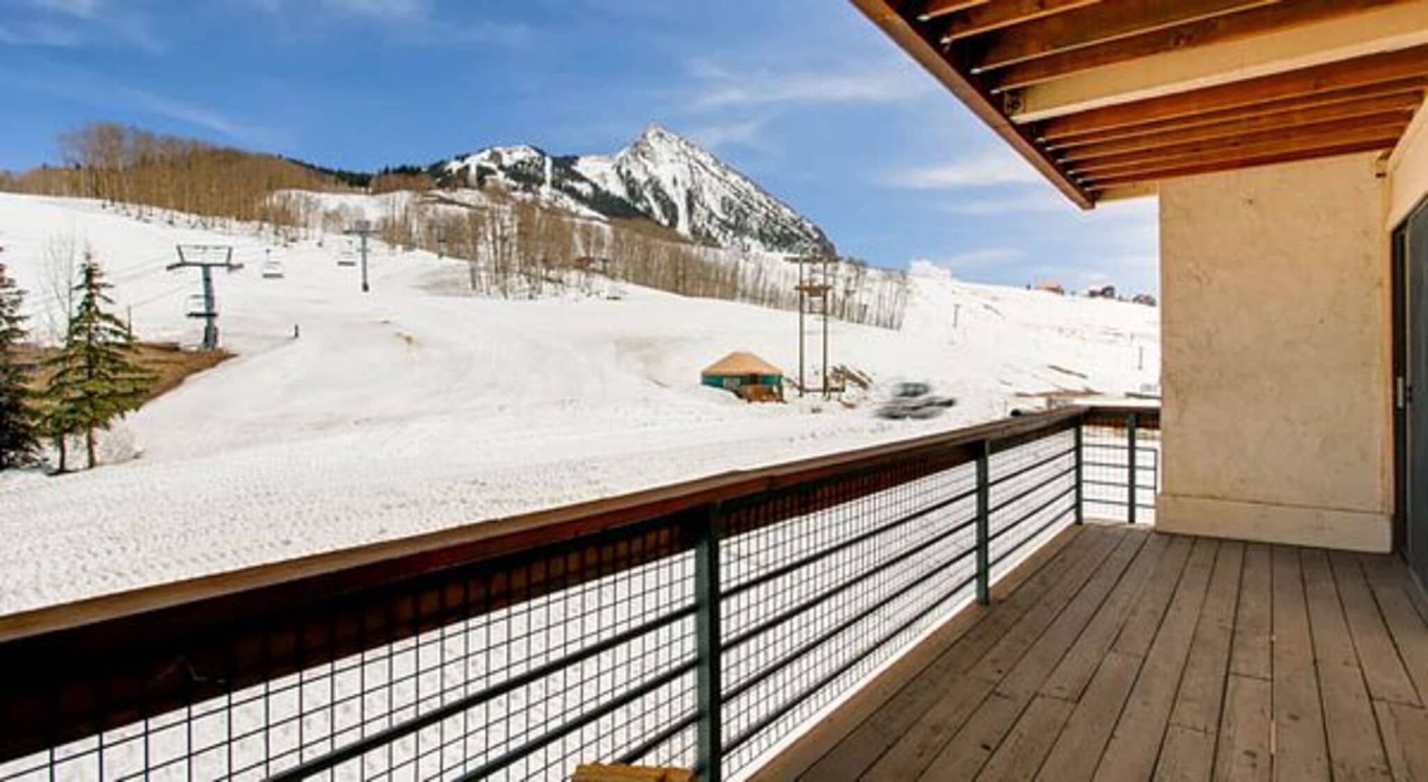 A wooden deck with a metal railing overlooking a snowy mountain slope with a ski lift and a small building.