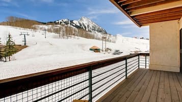 A wooden deck with a metal railing overlooking a snowy mountain slope with a ski lift and a small building.