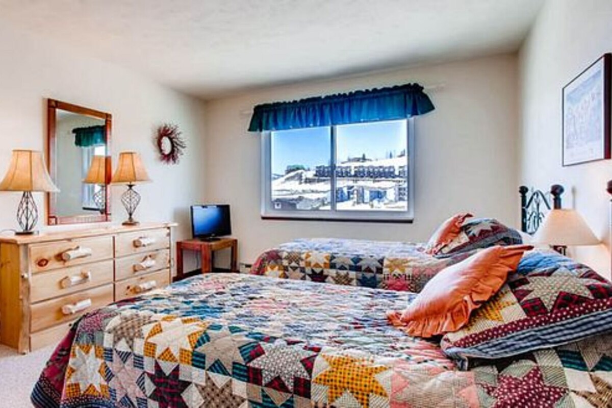 A bedroom with two beds, a dresser, a television, and a window with a view of buildings.