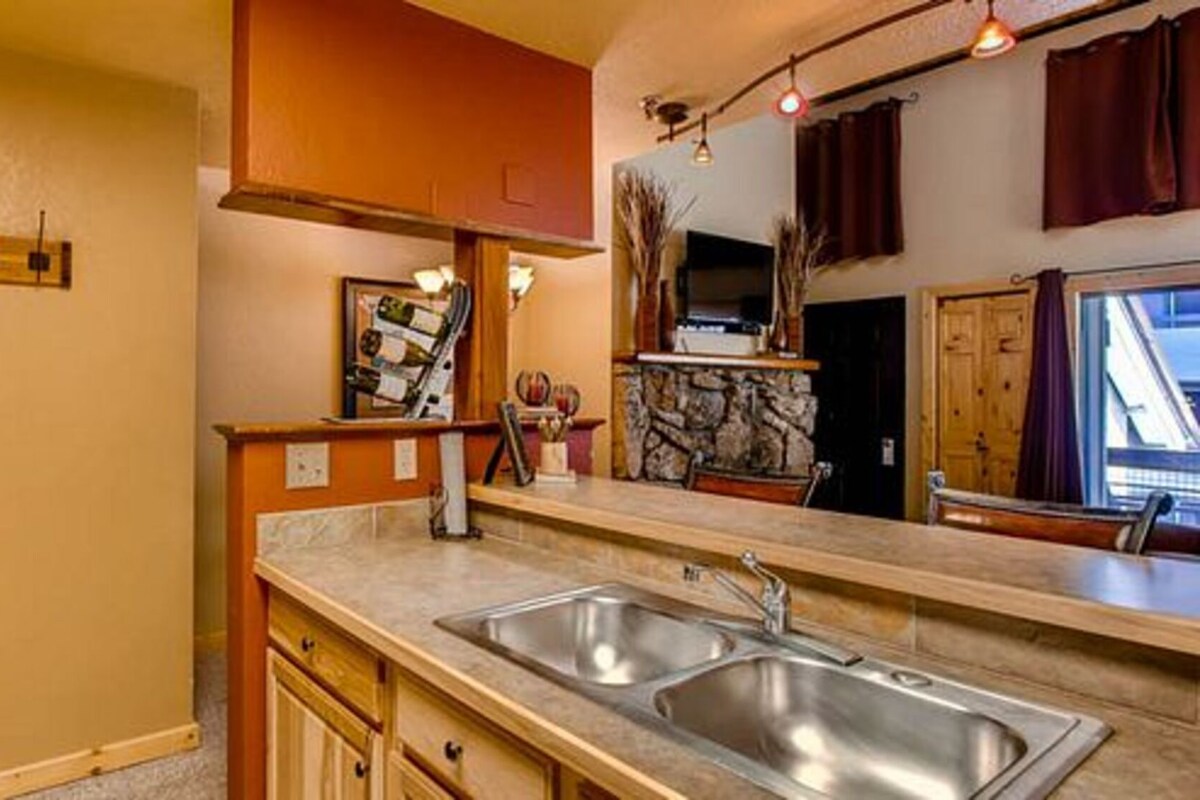 A kitchen with a double sink, wooden cabinets, a stone backsplash, and a window with curtains.
