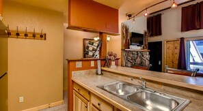 A kitchen with a double sink, wooden cabinets, a stone backsplash, and a window with curtains. - Crested Butte Mountain Resort Properties by Vail (Crested Butte)