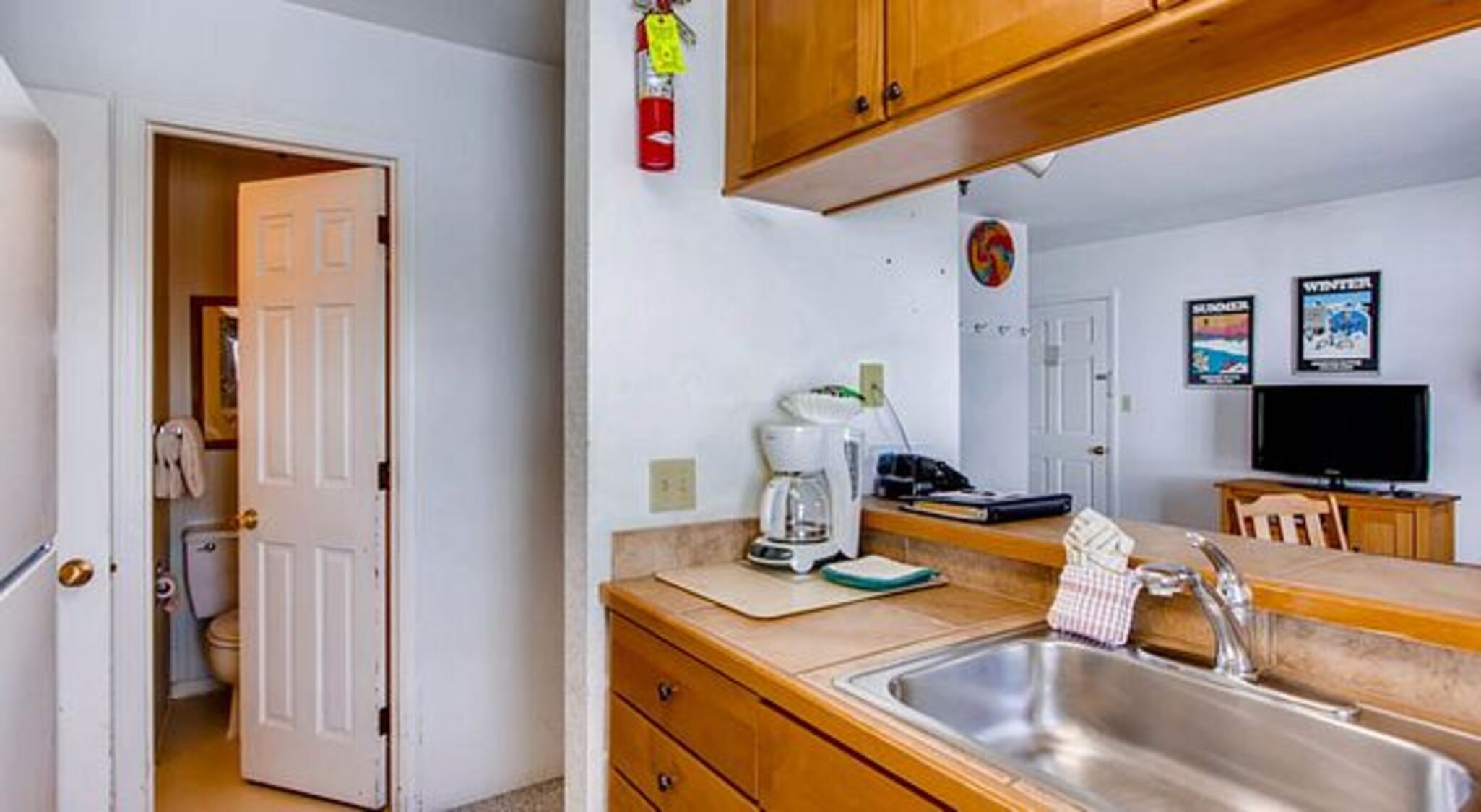 A compact kitchen with wooden cabinets, a coffee maker, a sink, and a television in the background.