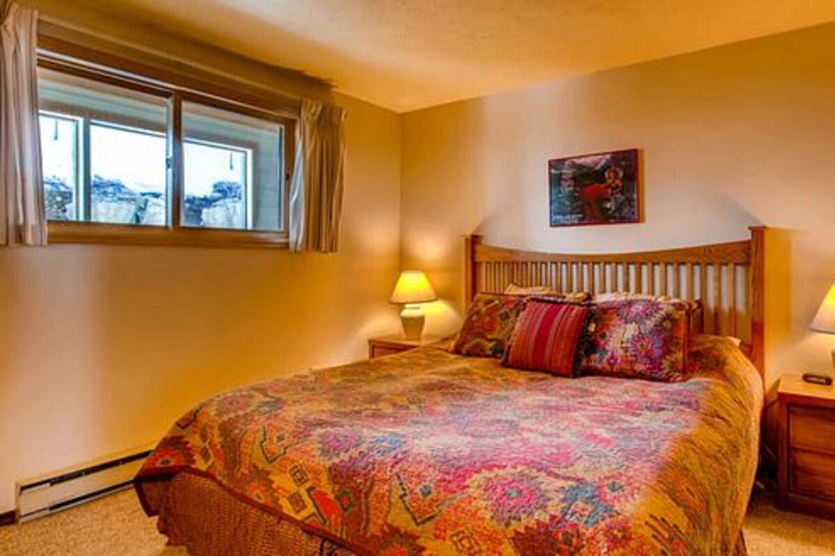 A bedroom with a large bed, two bedside lamps, a window with curtains, and a painting on the wall.