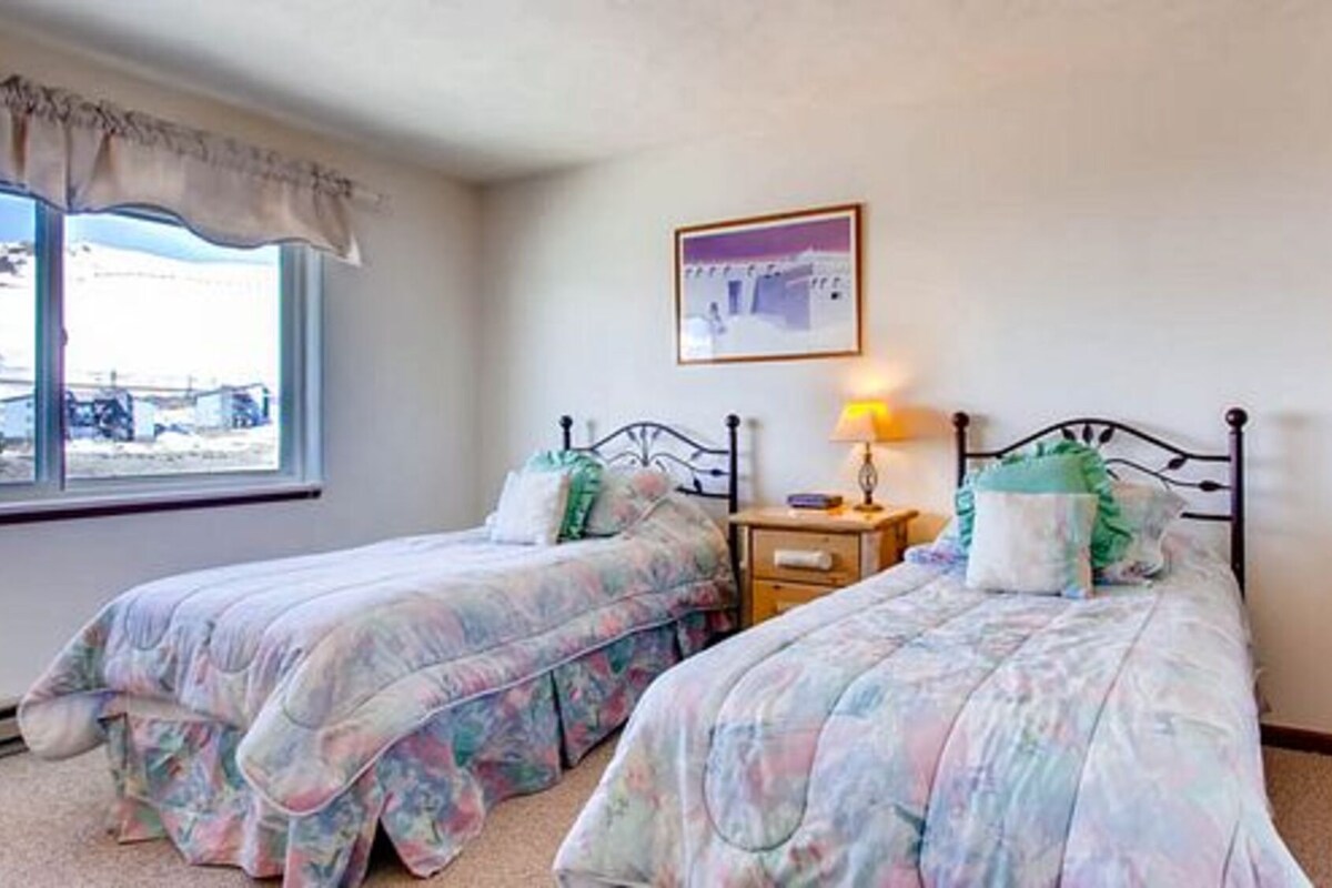A bedroom with two single beds, a nightstand with a lamp, a window with a view, and a cactus plant.